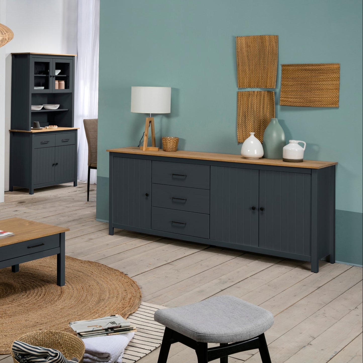 A staged living room furniture set featuring a dark gray sideboard with a light wood top, a matching TV stand, a coffee table, and a tall shelving unit, set against a teal wall and light wood floors.