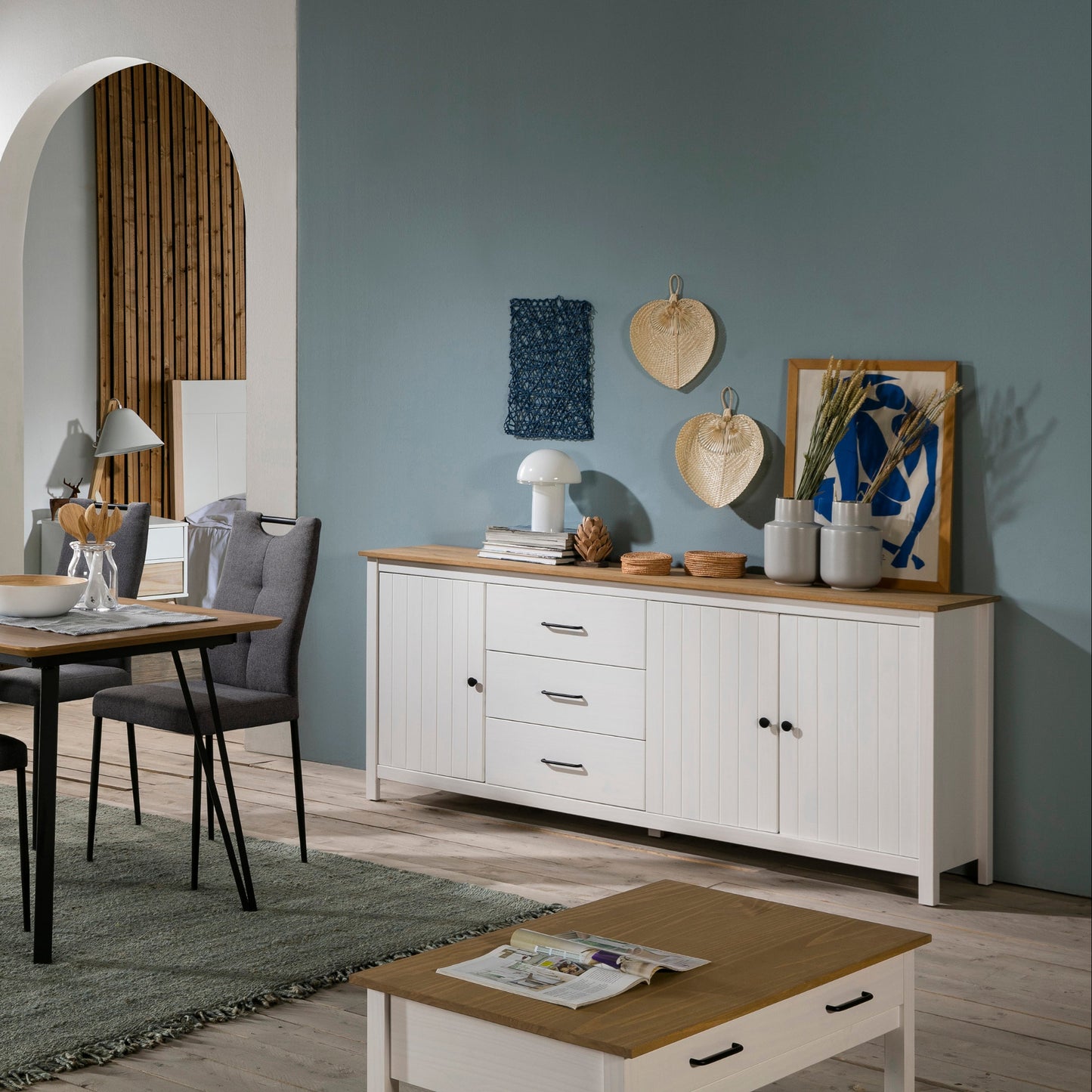 A styled dining room interior featuring a white dining table with six gray upholstered chairs, a matching white sideboard with a light wood top, and a white cabinet against a teal wall, with a light wood floor and rug.
