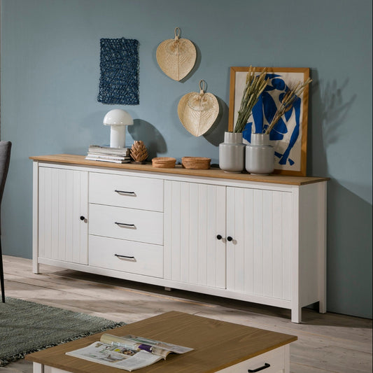 A styled dining room interior featuring a white dining table with six gray upholstered chairs, a matching white sideboard with a light wood top, and a white cabinet against a teal wall, with a light wood floor and rug.
