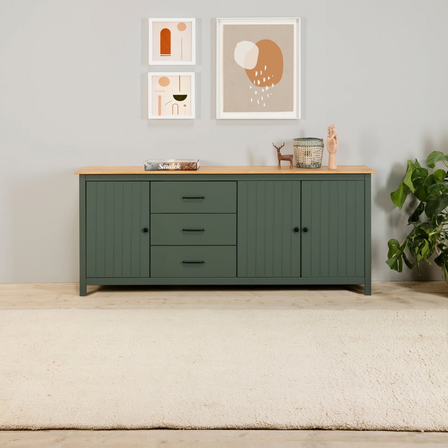 A modern sideboard cabinet in dark green with a light natural wood top, styled in a minimalist room setting with framed art, a plant, and a rug.