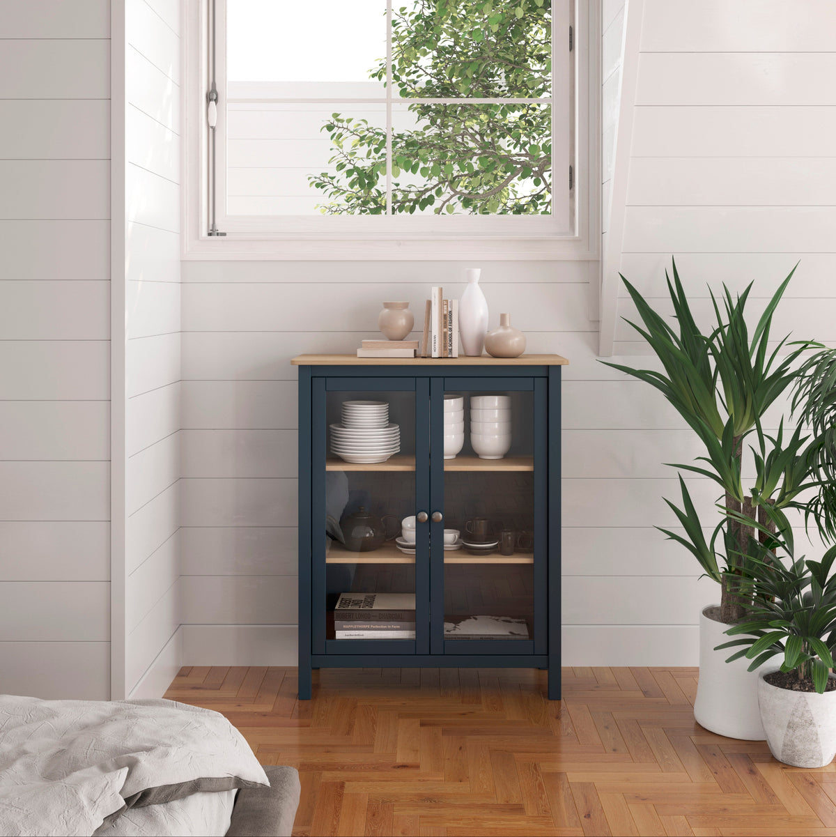 A navy blue glass-front cabinet with a light wood top stands in a bright room with white shiplap walls, a window with shutters, and a potted green plant nearby.