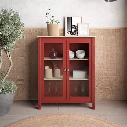A red wooden cabinet with glass doors and light wood shelves stands against a textured wall in a styled interior. The cabinet is decorated with small home decor items, framed art, and a potted plant, with two pendant lights and a woven rug visible in the room.