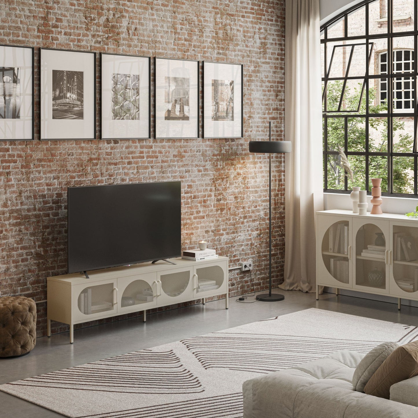 A modern living room featuring a white TV stand with arched glass-front doors, a black flat-screen TV, a black floor lamp, a brick wall with framed black-and-white prints, and a large window with sheer curtains.