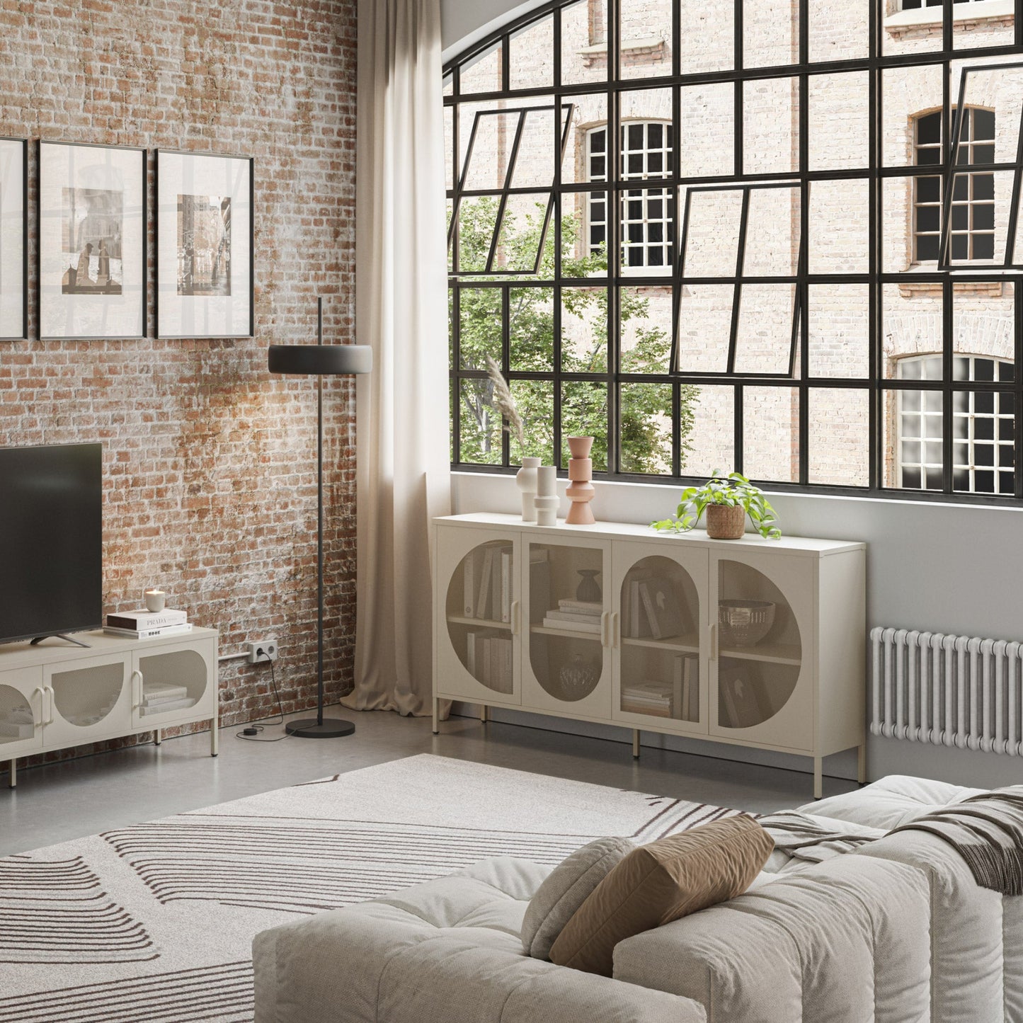 A modern living room featuring a cream sideboard cabinet with arched glass doors, styled in a bright room with a brick wall, large black-framed window, and a light-colored sofa.