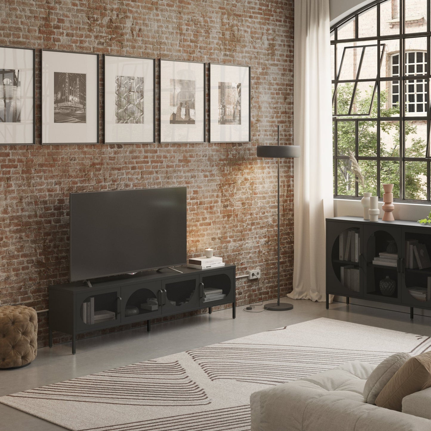 A modern living room scene featuring a black TV stand with a flat-screen television, set against a rustic brick wall with five framed black-and-white prints. A black floor lamp stands nearby, and a large window with white curtains is visible on the right.