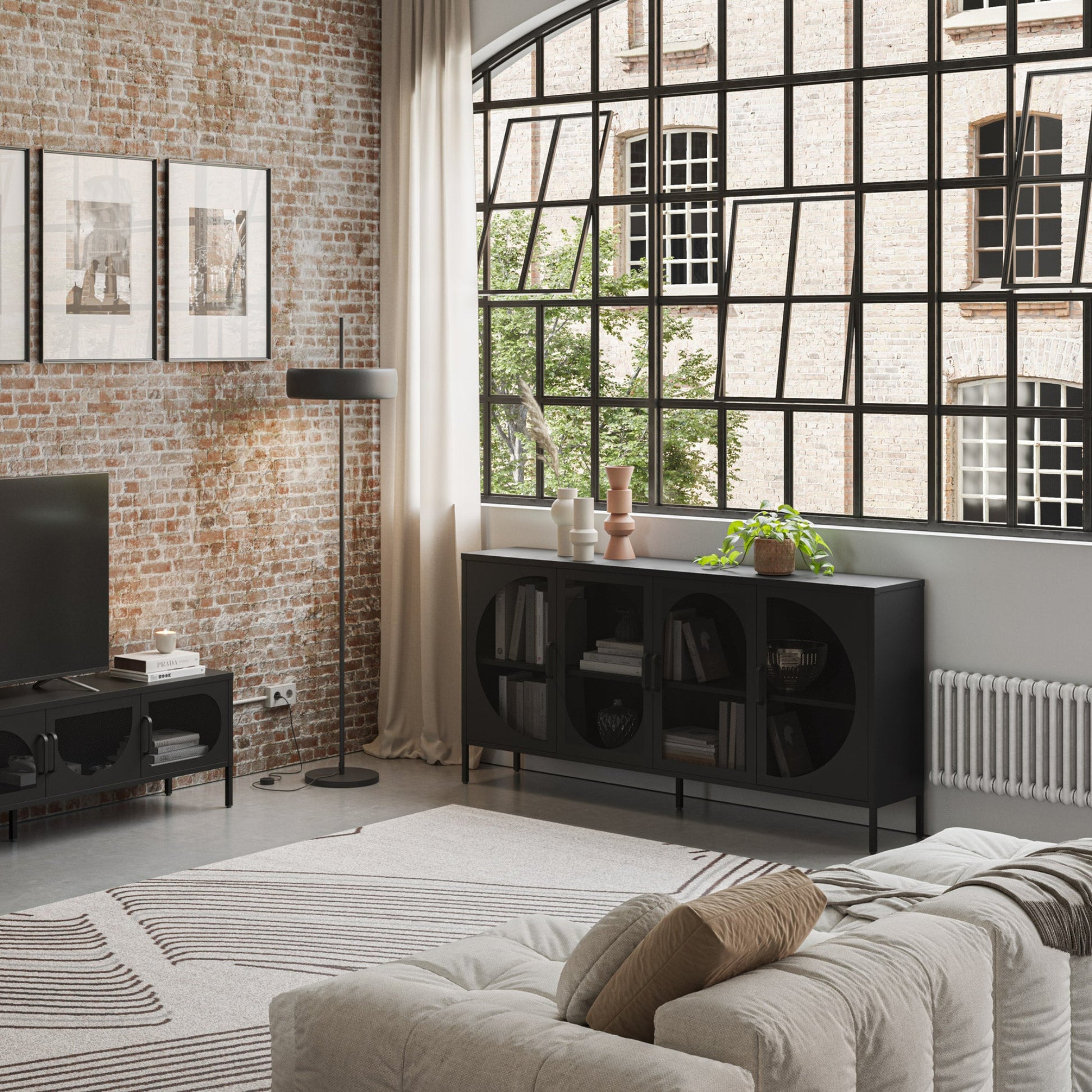 A modern living room interior featuring a black round-decorated sideboard cabinet with circular shelving, styled in an industrial loft setting with a brick wall, large multi-pane window, and a beige sofa in the foreground.