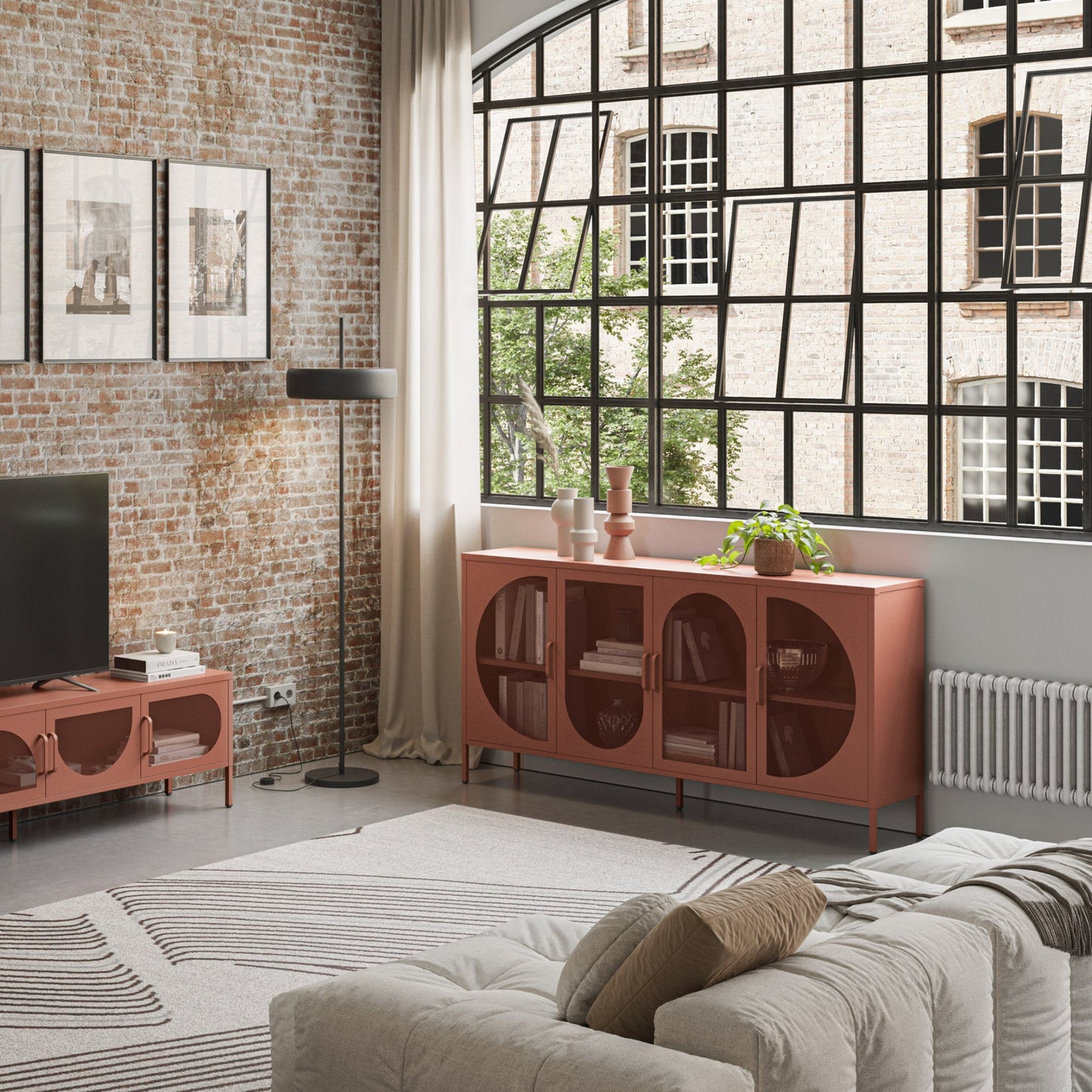 A modern living room featuring a terracotta-colored wooden sideboard cabinet with arched door panels, styled in an industrial loft setting with a brick wall, large black-framed window, and a matching sideboard under the window.
