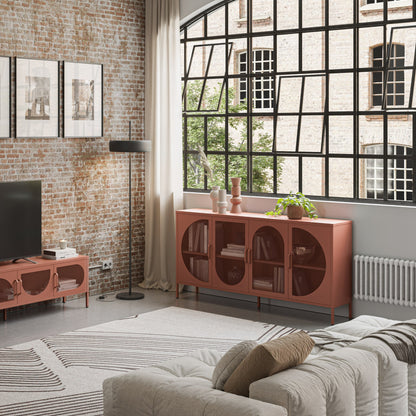 A modern living room featuring a terracotta-colored wooden sideboard cabinet with arched door panels, styled in an industrial loft setting with a brick wall, large black-framed window, and a matching sideboard under the window.