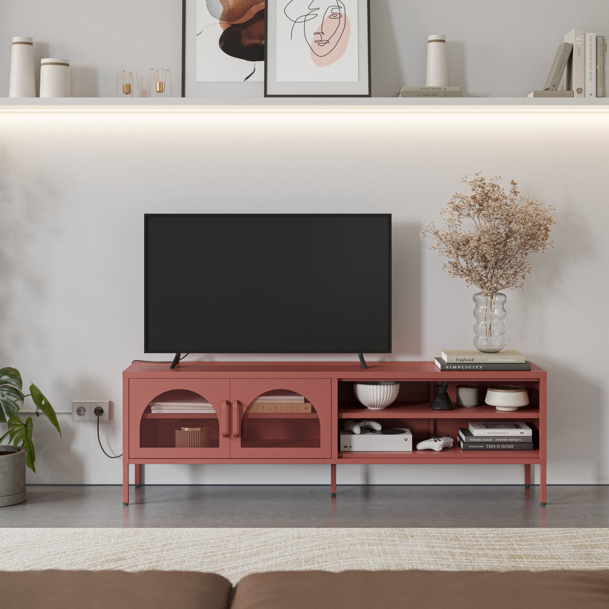 A modern terracotta red TV stand console in a living room setting, styled with a black flat-screen TV, books, bowls, and a vase of dried flowers against a white wall.