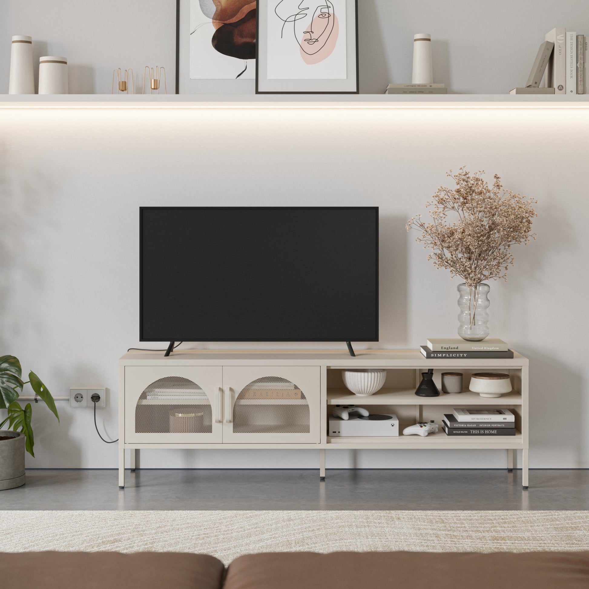 A modern light beige TV stand console with open shelves and arched cabinet doors, styled in a living room setting with a black flat-screen TV, decorative books, a vase of dried flowers, and wall shelves above.