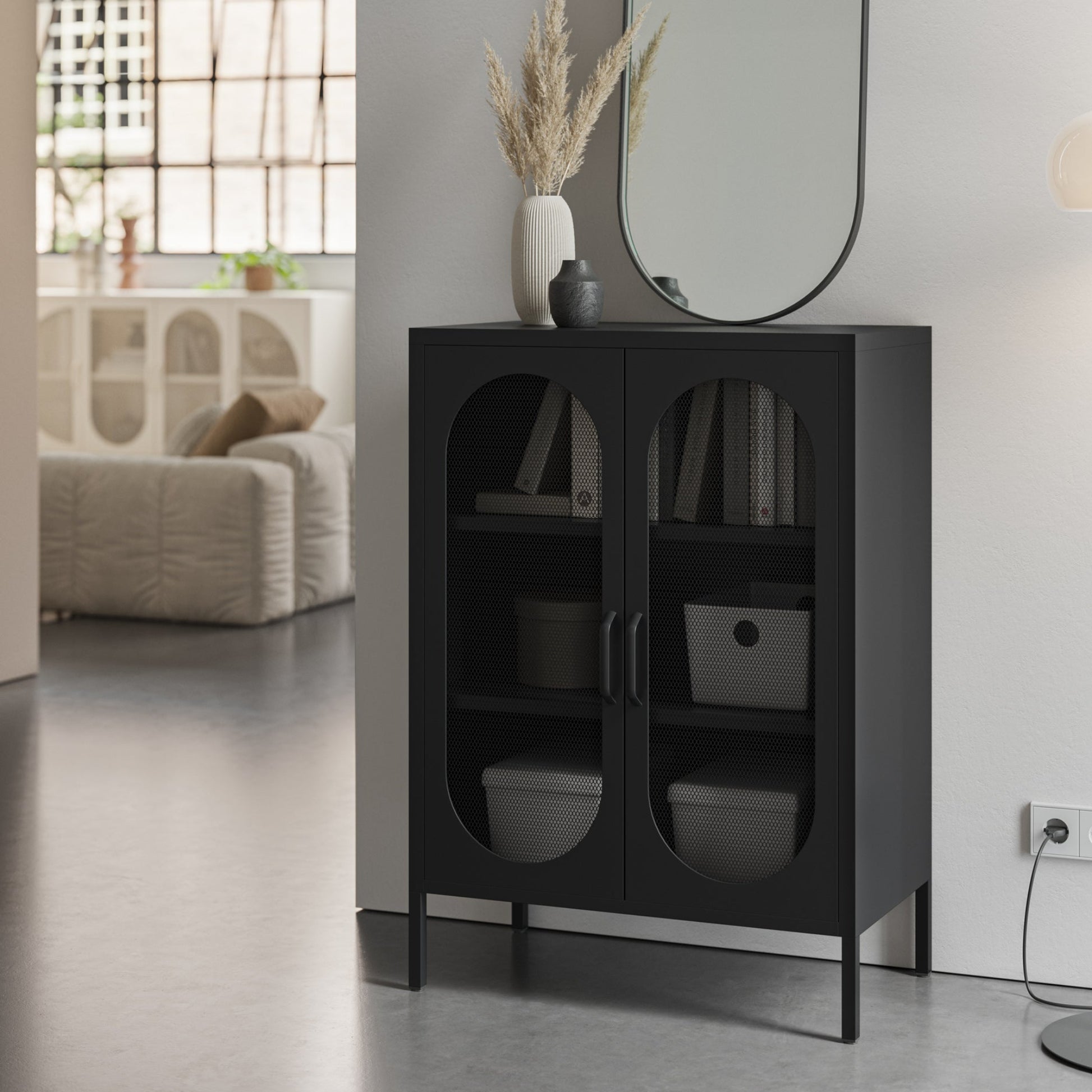 A modern black storage cabinet with two arched glass doors stands in a minimalist room setting next to a white wall, holding books and storage bins.