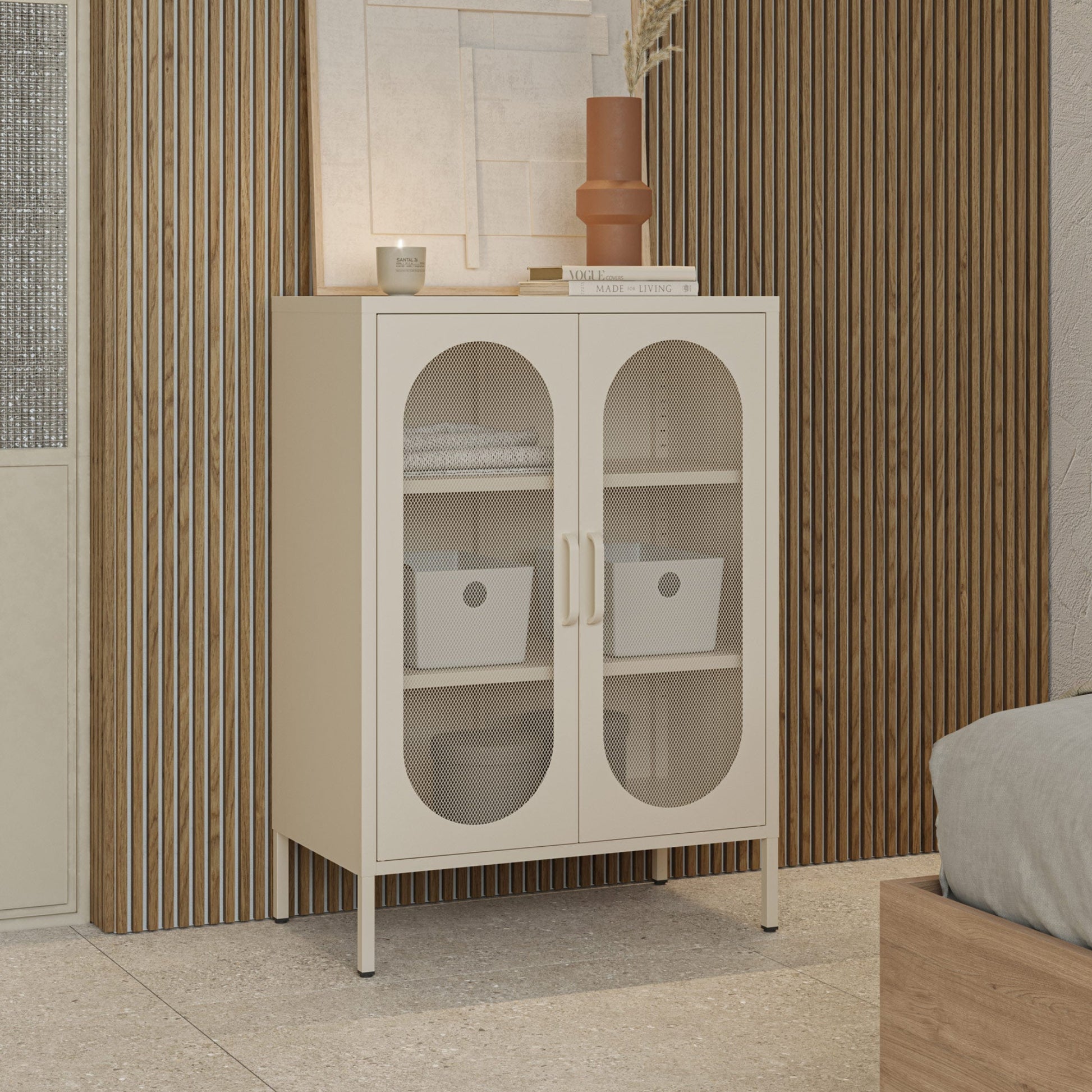 A small cream-colored storage cabinet with two arched glass doors stands in a bedroom or living space against a slatted wood wall. The cabinet has two light wood handles, four legs, and open shelves inside with folded towels, storage bins, and a vase on top.