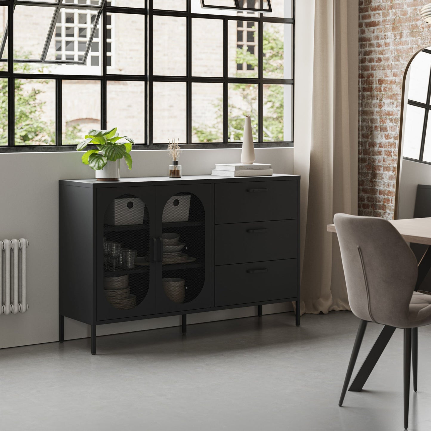 A modern black sideboard cabinet with glass-front doors and three drawers, styled in a contemporary loft room with a brick wall, large window, and a gray upholstered chair.