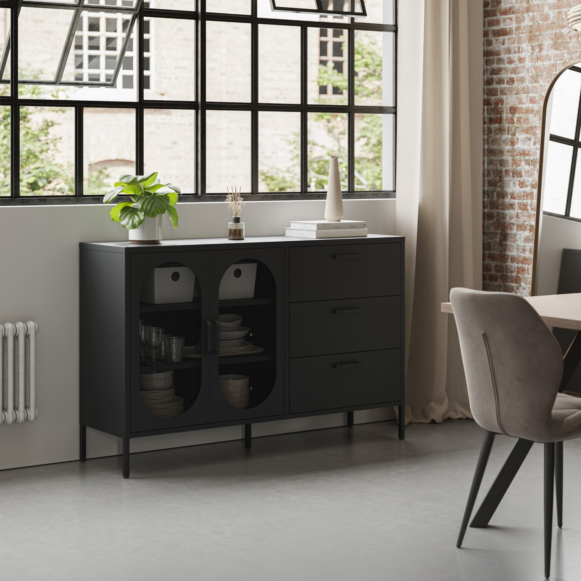 A modern black sideboard cabinet with glass-front doors and three drawers, styled in a contemporary loft room with a brick wall, large window, and a gray upholstered chair.