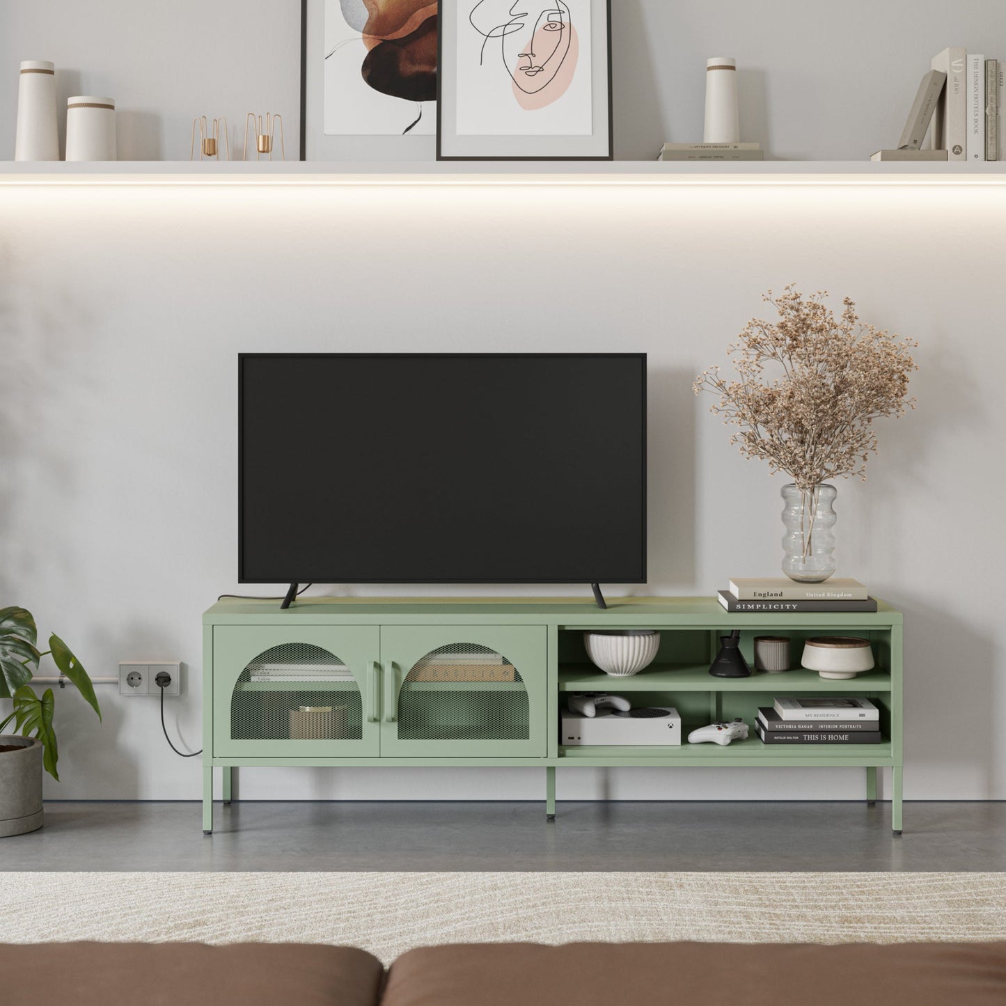 A light sage green TV stand console with glass-front cabinets and open shelving, styled in a modern living room setting with a black flat-screen TV, decorative bowls, books, and a vase of dried flowers.
