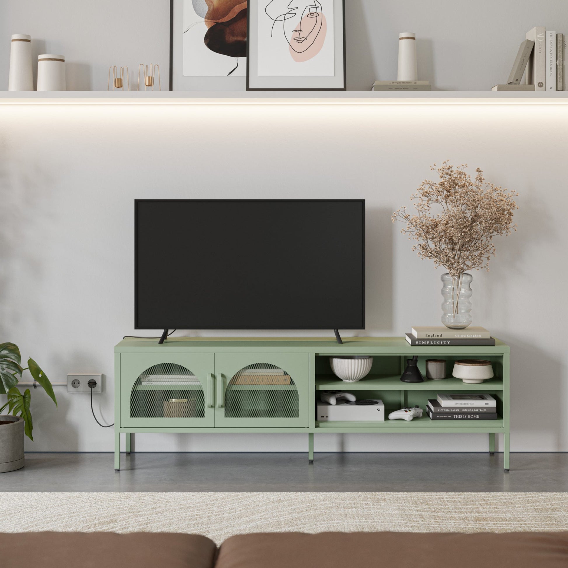 A light sage green TV stand console with glass-front cabinets and open shelving, styled in a modern living room setting with a black flat-screen TV, decorative bowls, books, and a vase of dried flowers.