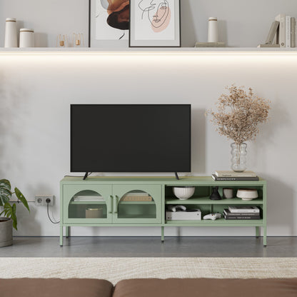 A light sage green TV stand console with glass-front cabinets and open shelving, styled in a modern living room setting with a black flat-screen TV, decorative bowls, books, and a vase of dried flowers.