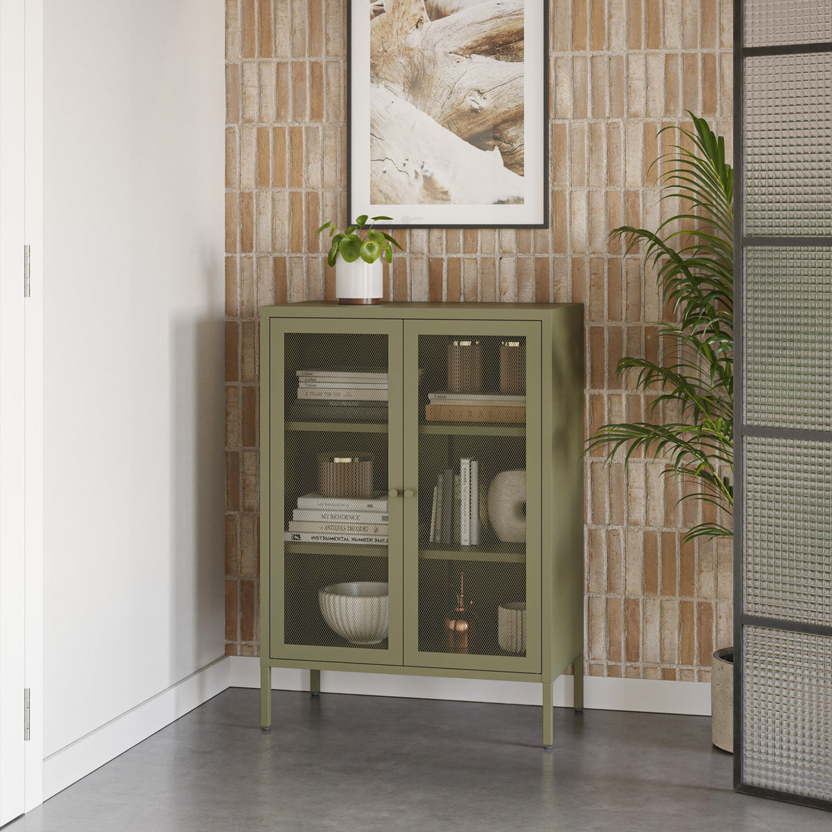An olive green glass-front cabinet with legs stands against a textured light brown brick wall, styled with books, bowls, and a small potted plant. A framed print hangs above, and a tall plant and black screen are visible in the room.