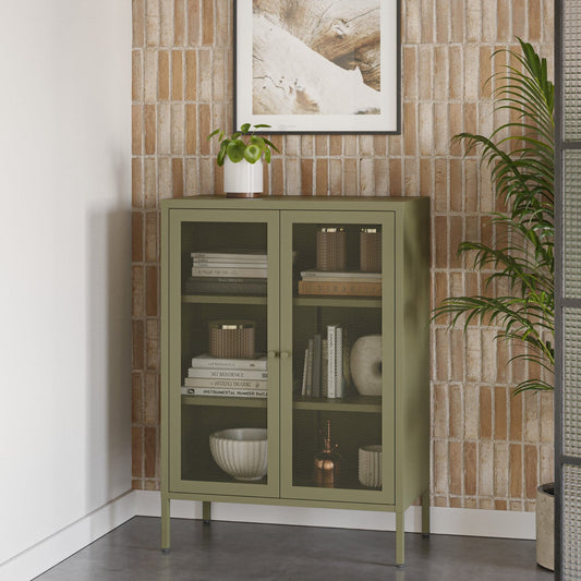 An olive green glass-front cabinet with legs stands against a textured light brown brick wall, styled with books, bowls, and a small potted plant. A framed print hangs above, and a tall plant and black screen are visible in the room.