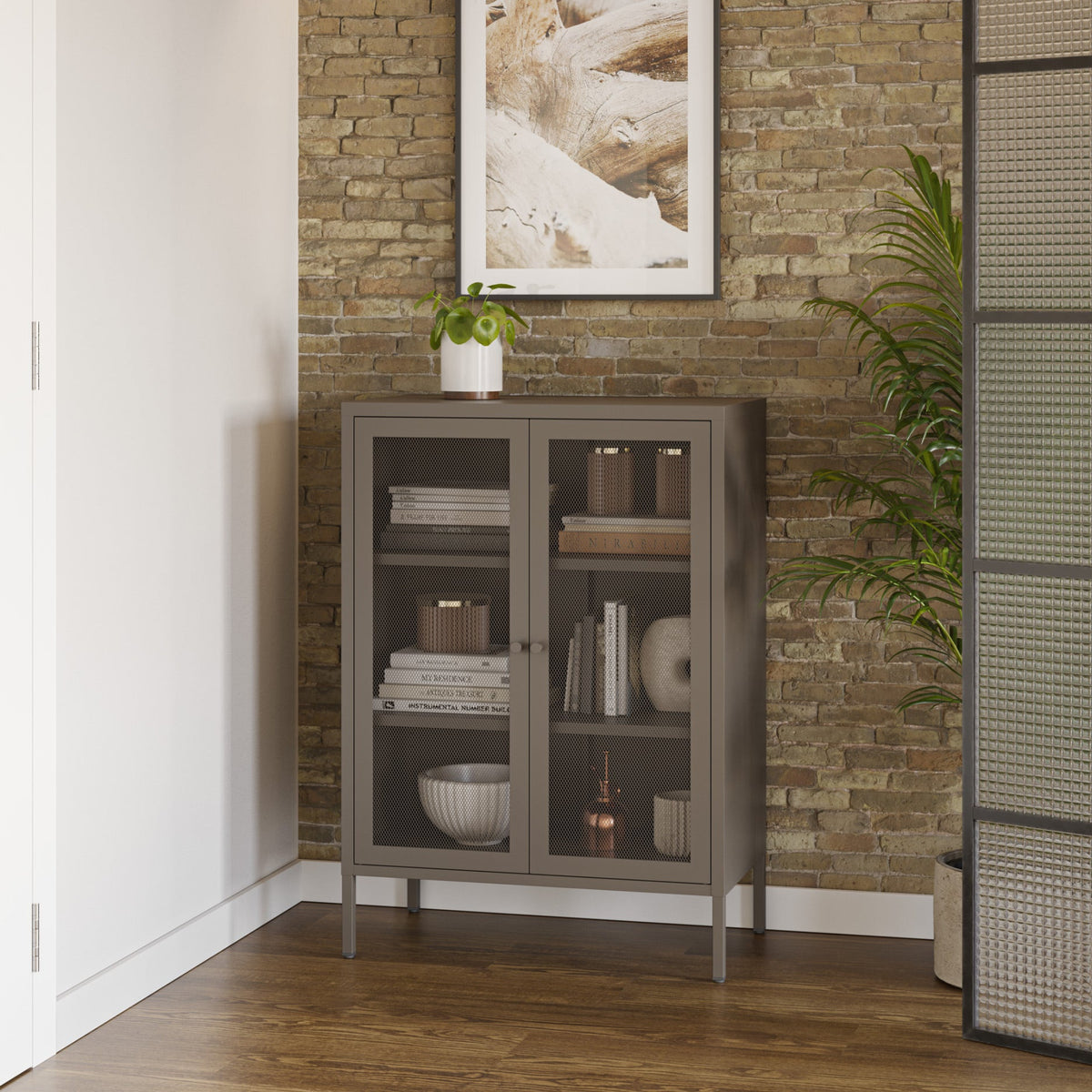 A tall gray cabinet with glass doors stands against a rustic brick wall in a styled interior. A framed abstract print hangs above it, with a small potted plant on top and a tall green plant to the right.