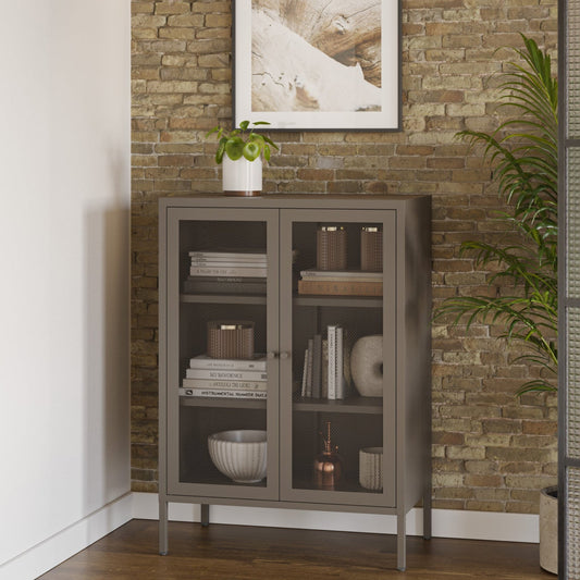 A tall gray cabinet with glass doors stands against a rustic brick wall in a styled interior. A framed abstract print hangs above it, with a small potted plant on top and a tall green plant to the right.
