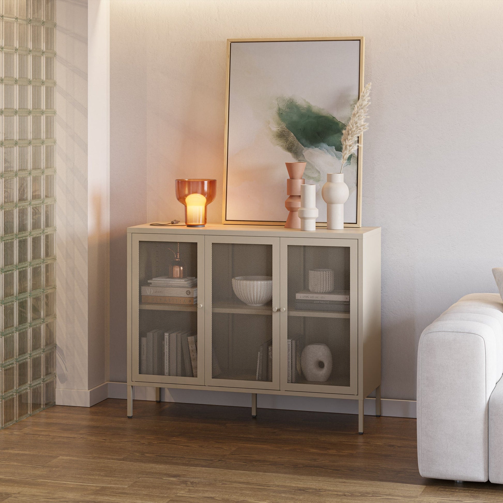A light beige wood cabinet with three glass doors stands against a neutral wall in a styled room. Decorative objects, books, and a framed print sit on top of the cabinet, which is placed in a living space next to a white sofa and a glass block screen.