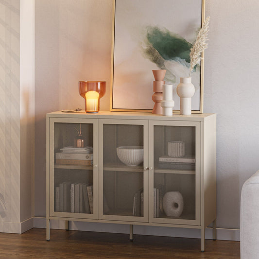 A light beige wood cabinet with three glass doors stands against a neutral wall in a styled room. Decorative objects, books, and a framed print sit on top of the cabinet, which is placed in a living space next to a white sofa and a glass block screen.