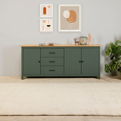 A modern sideboard cabinet in dark green with a light natural wood top, styled in a minimalist room with framed art, a plant, and a cream rug.
