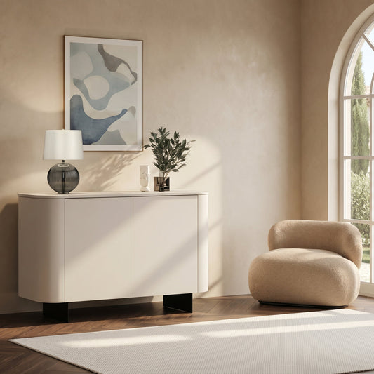 A modern white sideboard or credenza in a styled interior room, topped with a table lamp and a small plant. The room has warm beige walls, a large arched window, a beige armchair, and a light rug.