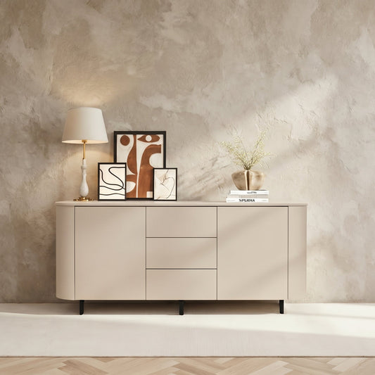 A modern sideboard cabinet in a warm cream-beige finish, styled in a minimalist room with a textured plaster wall, a white rug, a table lamp, framed art, and a small vase.