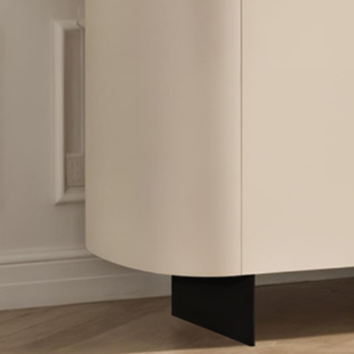 A close-up of a modern sideboard cabinet with curved cream-colored doors and a black triangular metal base, set against a white paneled wall and light wood flooring.