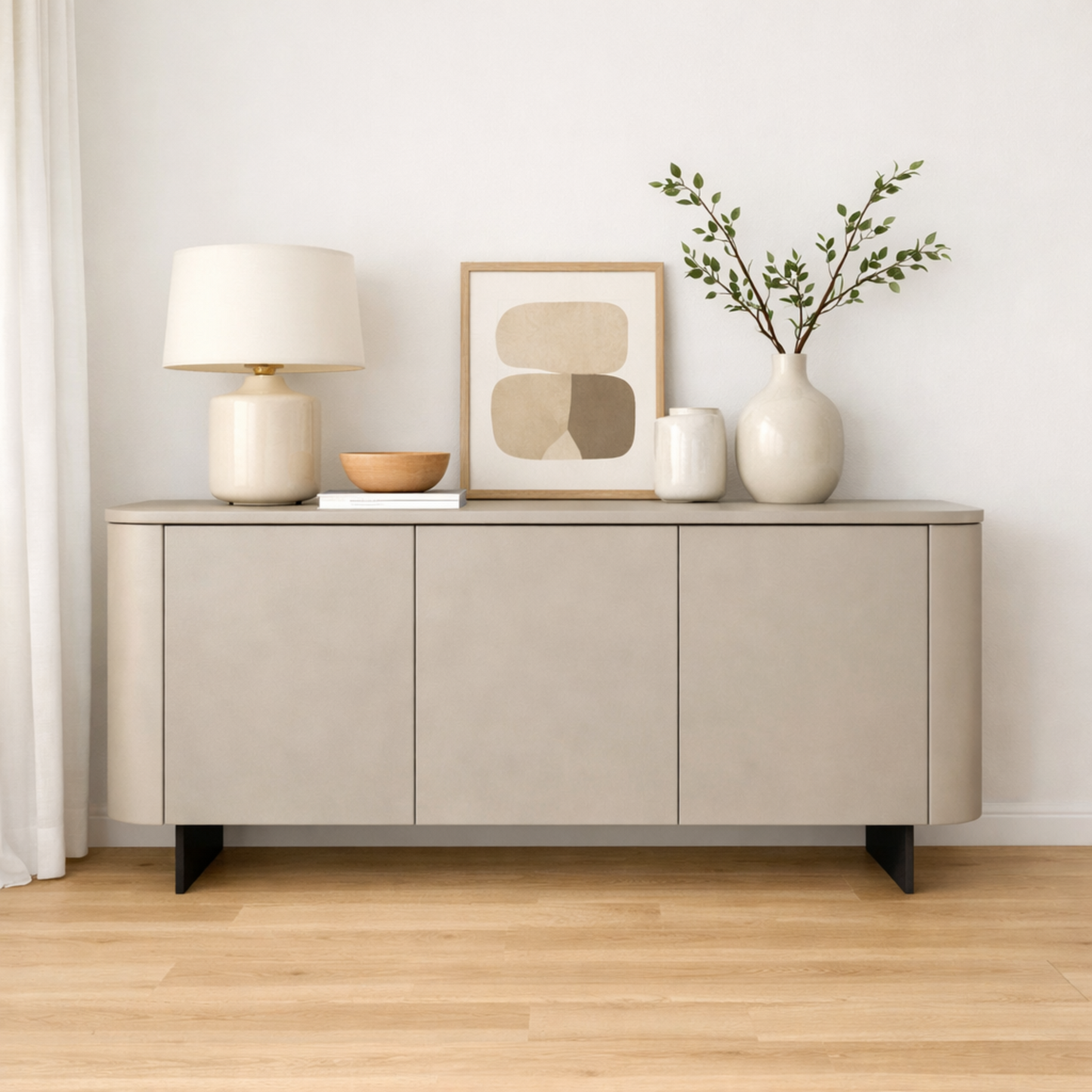 A modern light beige sideboard or credenza with black legs, styled with a table lamp, framed abstract art, a small bowl, a vase, and greenery against a white wall and light wood floor.