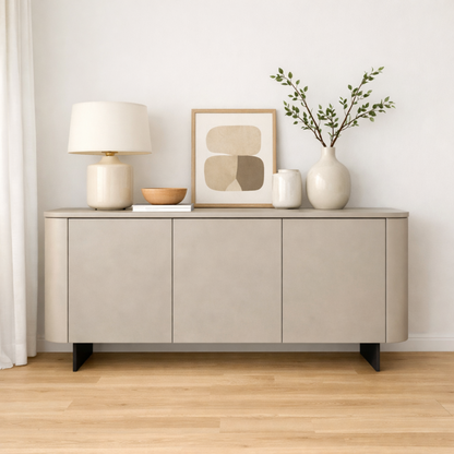 A modern light beige sideboard or credenza with black legs, styled with a table lamp, framed abstract art, a small bowl, a vase, and greenery against a white wall and light wood floor.