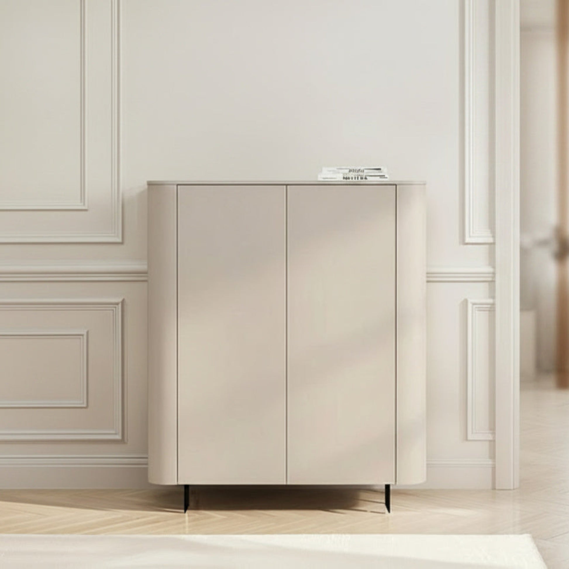 A modern, minimalist two-door cabinet in a warm beige tone, styled in a bright, elegant room with classic white paneled walls and light wood flooring.