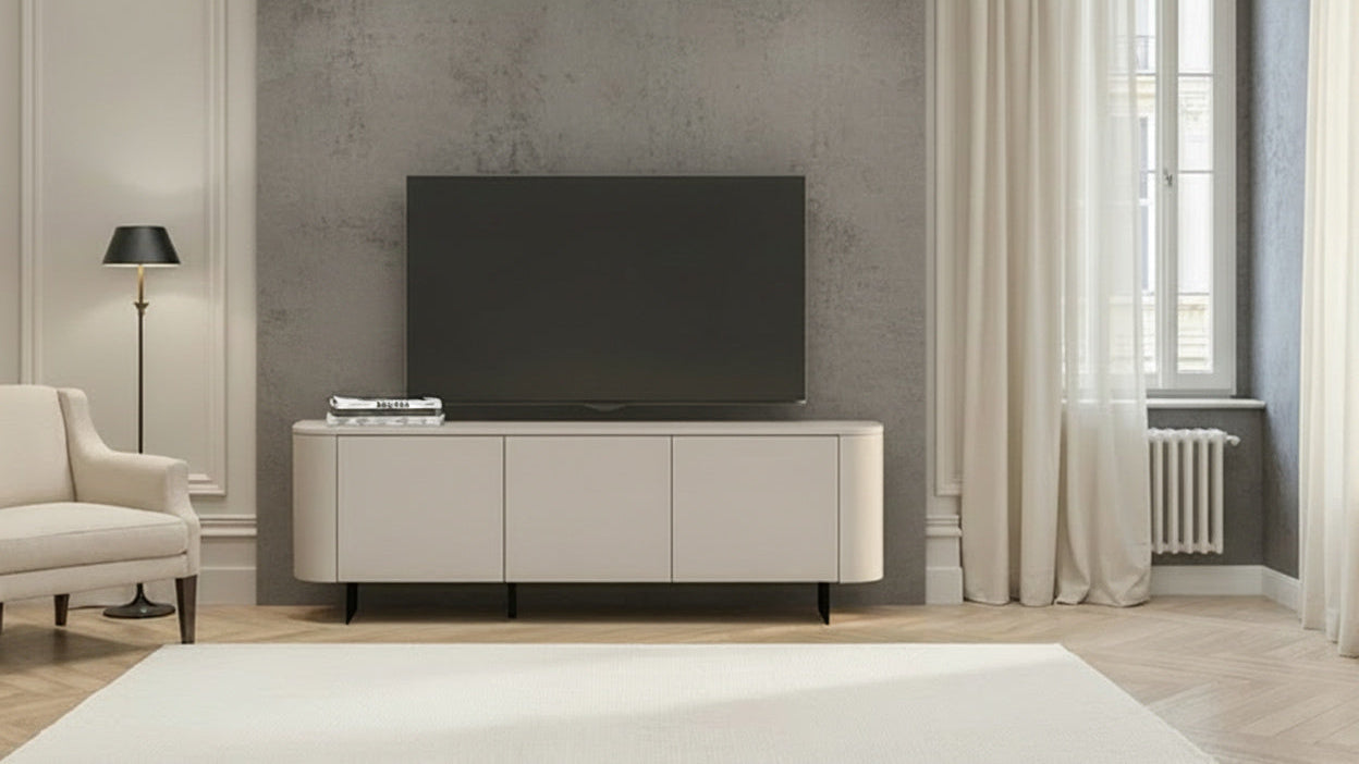 A modern beige media console with a black flat-screen TV on top, set against a textured gray wall in a styled living room with a white armchair, floor lamp, and curtains.