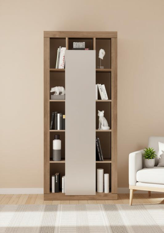 A tall wooden bookcase with sliding doors in a warm medium brown finish, styled with books, white decorative figurines, and cylindrical vases, set against a beige wall in a modern living room next to a white armchair.