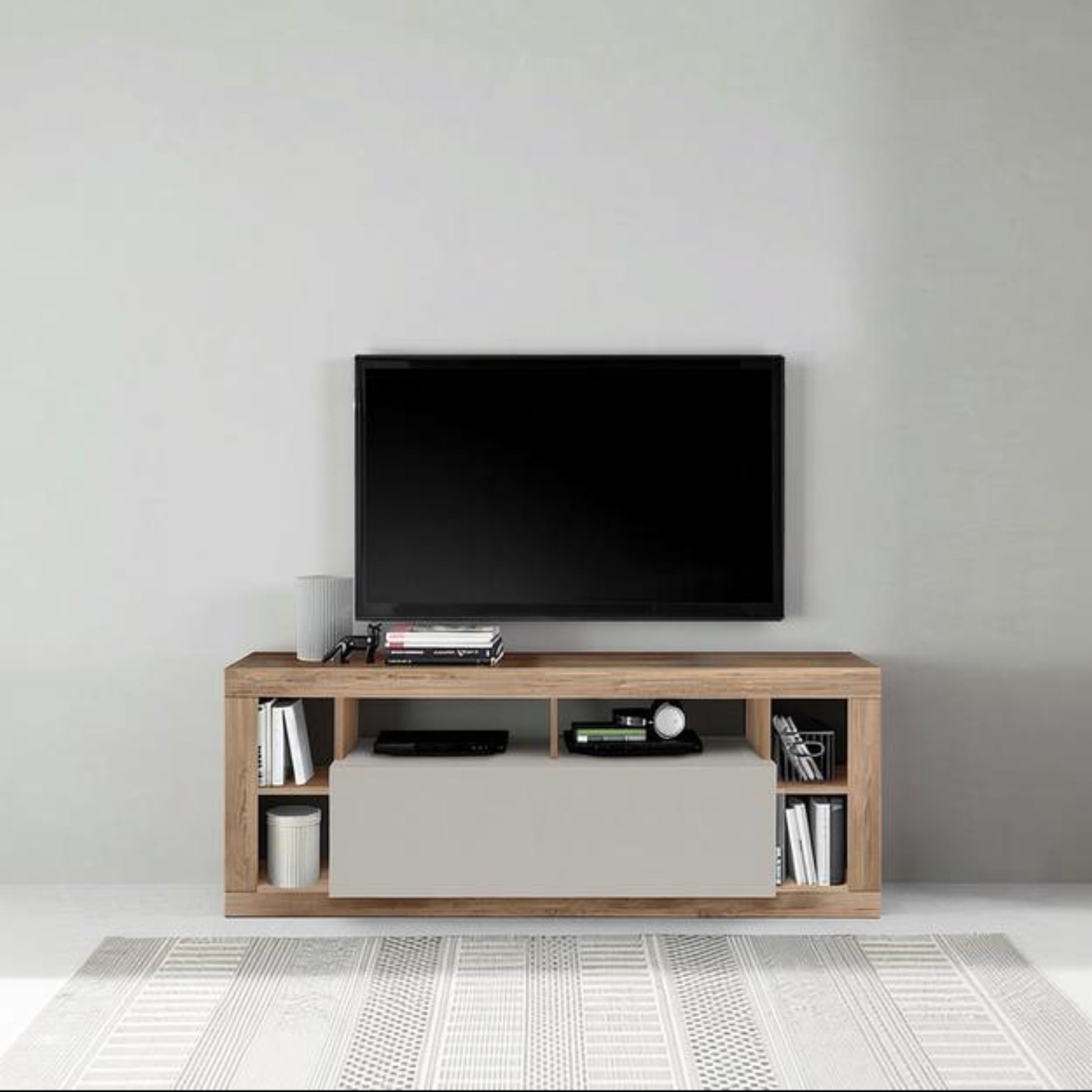A modern wooden TV stand console with a light oak finish and a central cream drawer, styled with a black flat-screen TV, electronic devices, and books, set against a minimalist gray wall and white patterned rug.
